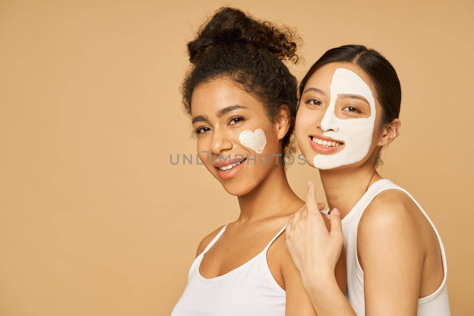 Two young female friends having fun, smiling at camera while posing with facial masks on isolated over beige background by friendsstock