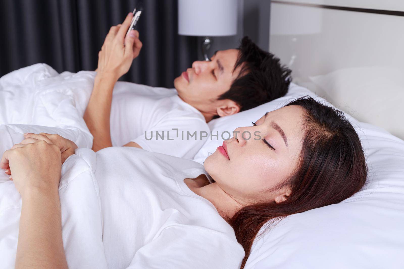 woman in bed with her sleeping and her husband texting on smartphone in bedroom by geargodz