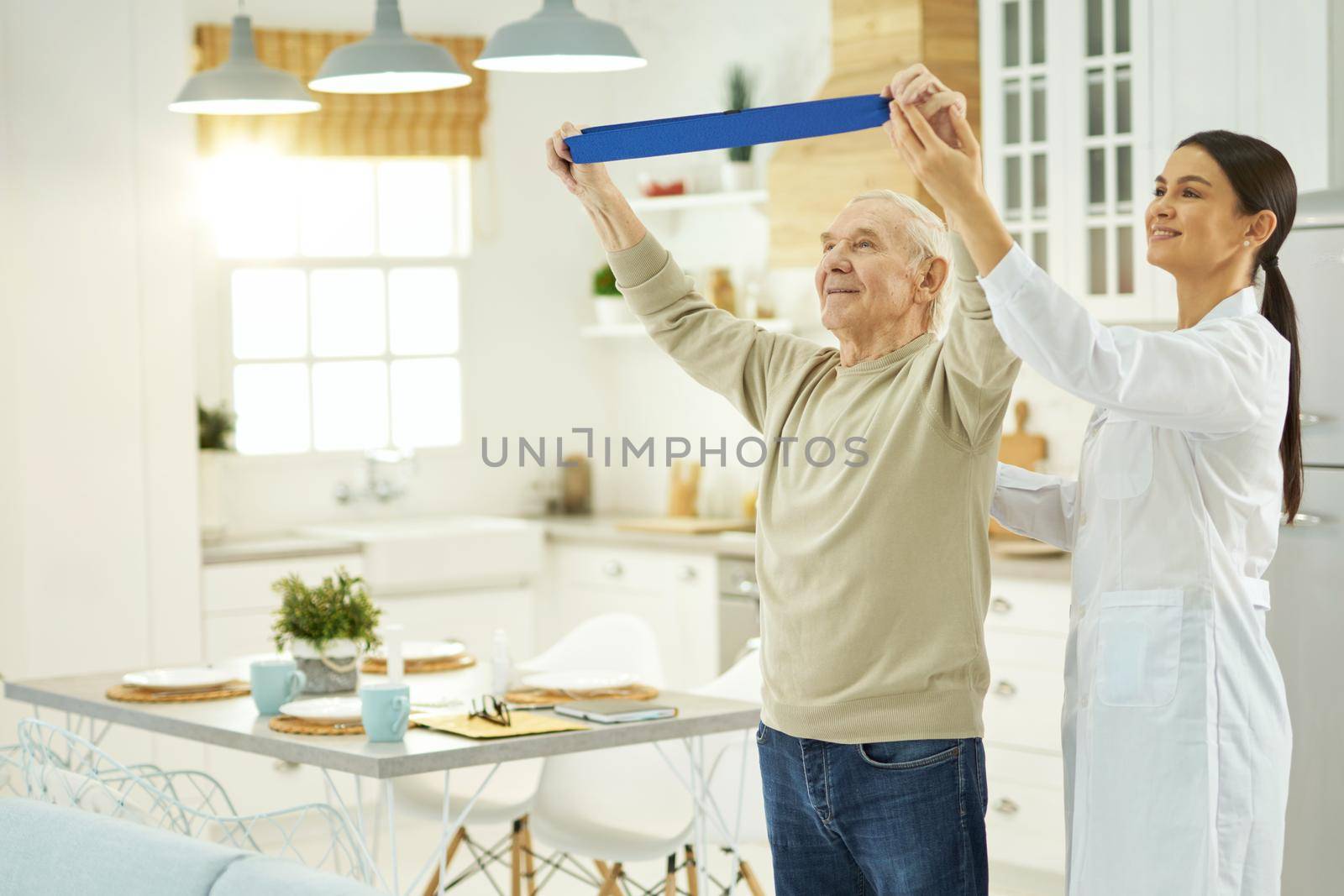 Aged man raising his hands up and doing exercises with fitness rubber band, female doctor helping him