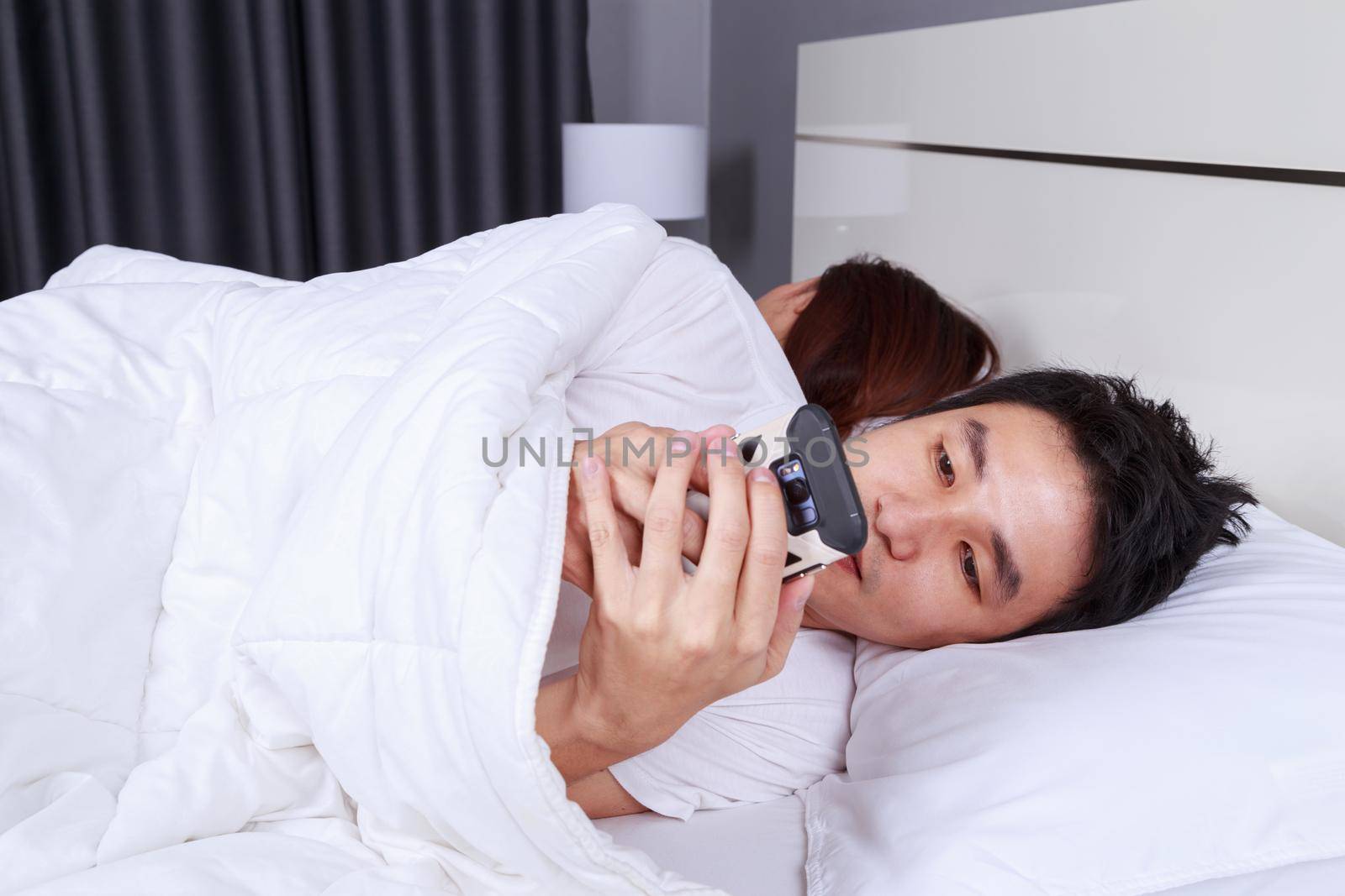 man using his mobile phone in bed while his wife is sleeping next to him by geargodz