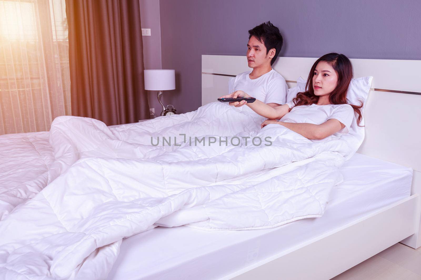 young couple lying on a bed with remote control and watching television in bedroom by geargodz