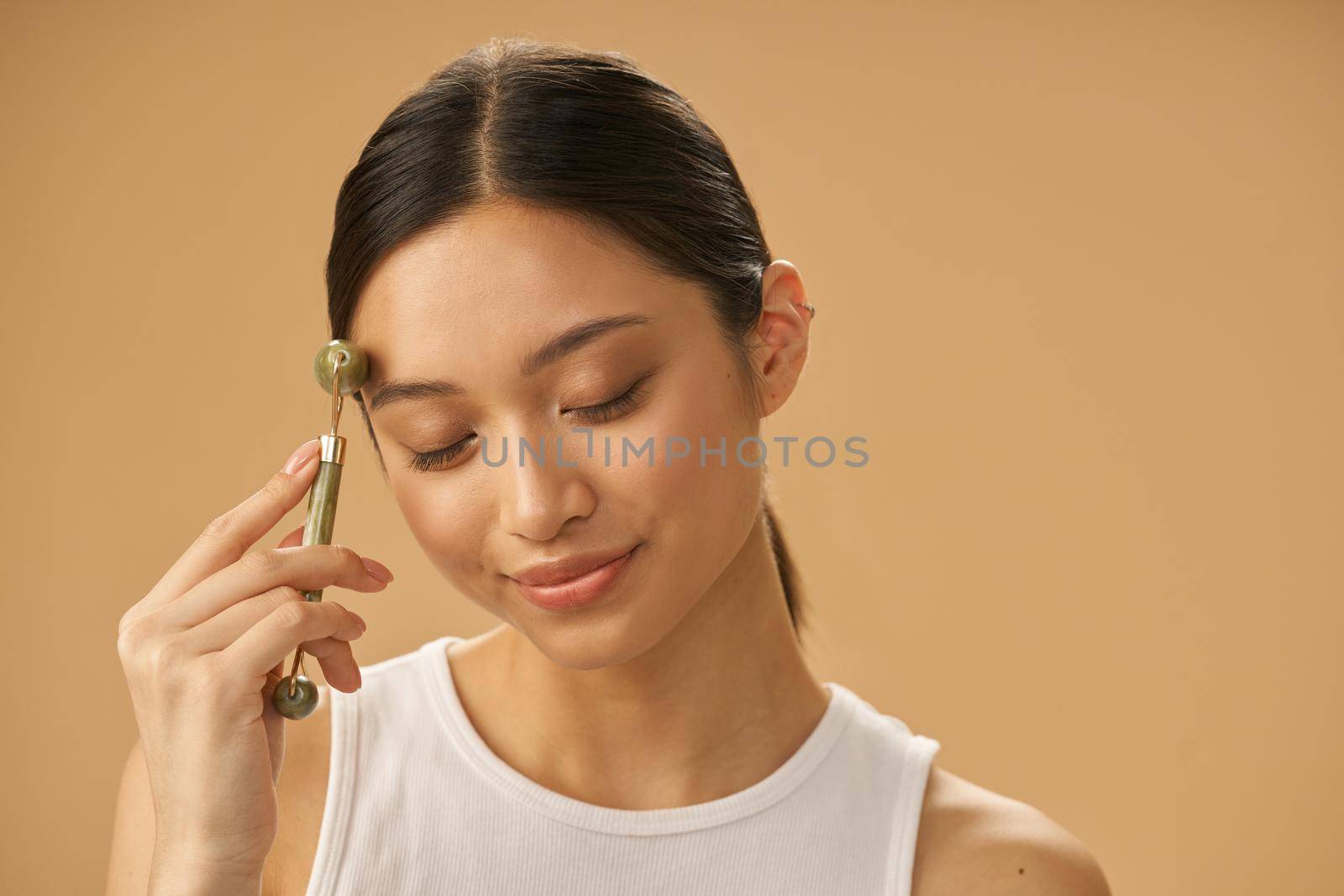 Relaxed young woman using jade roller for massaging her face, posing with eyes closed isolated over beige background. Skincare concept