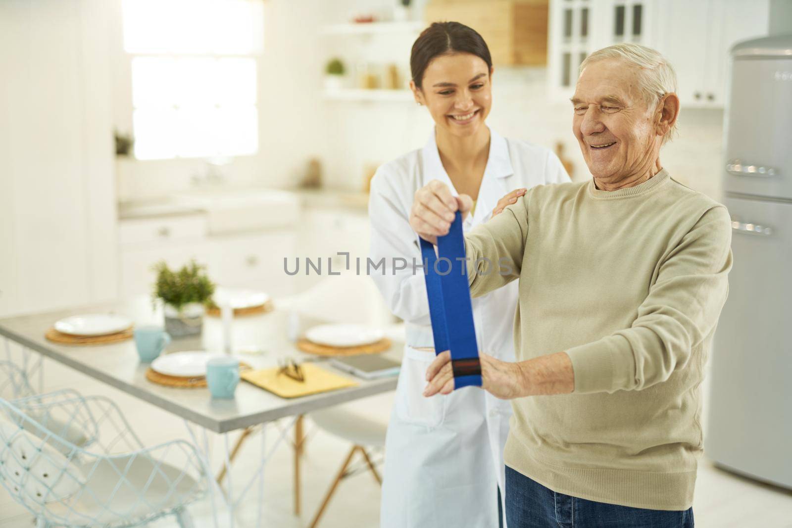 Smiling pensioner doing exercises and holding a fitness elastic band in hands while talking to nurse