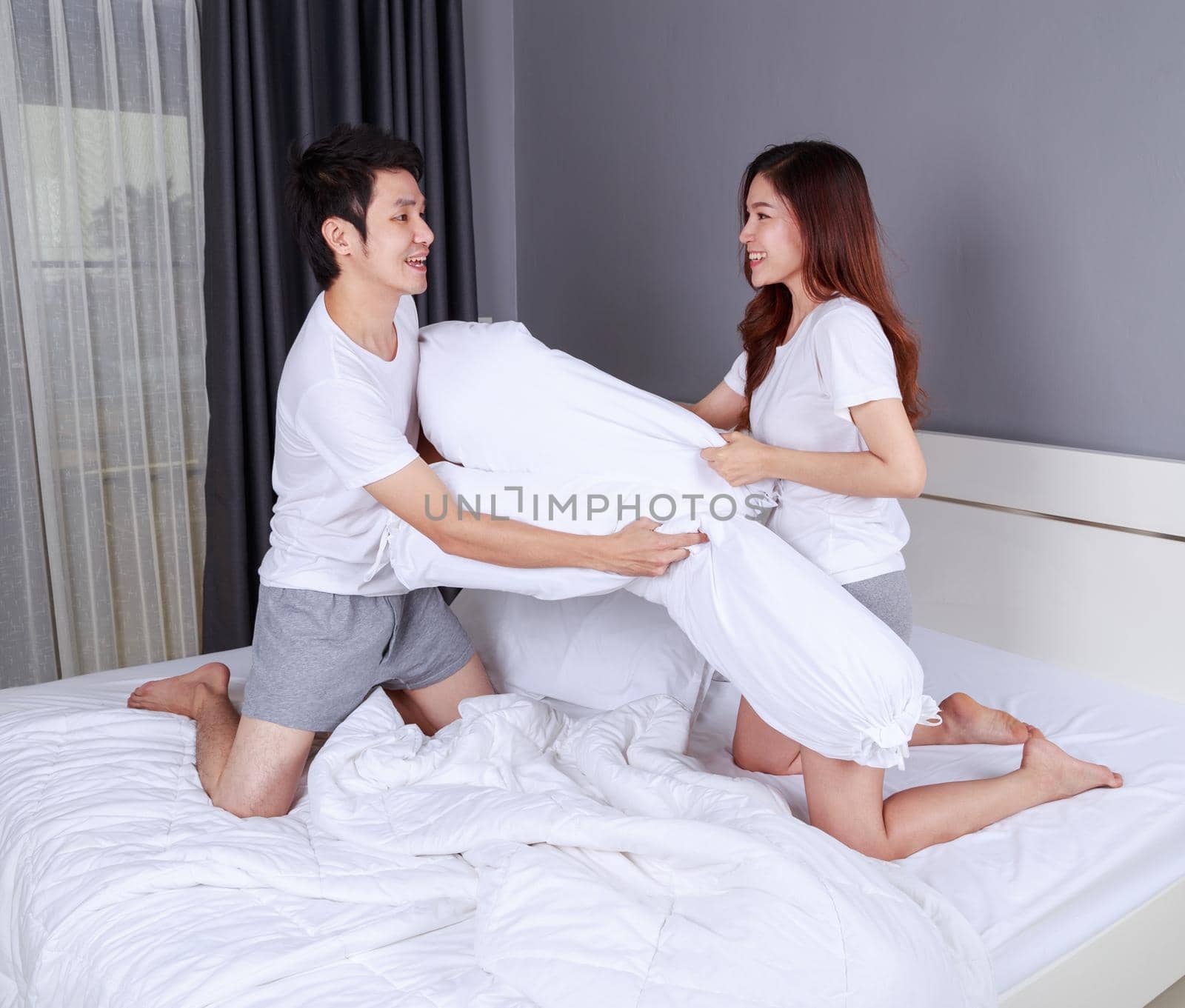 cheerful young couple having a bolster pillow fight on bed in the bedroom