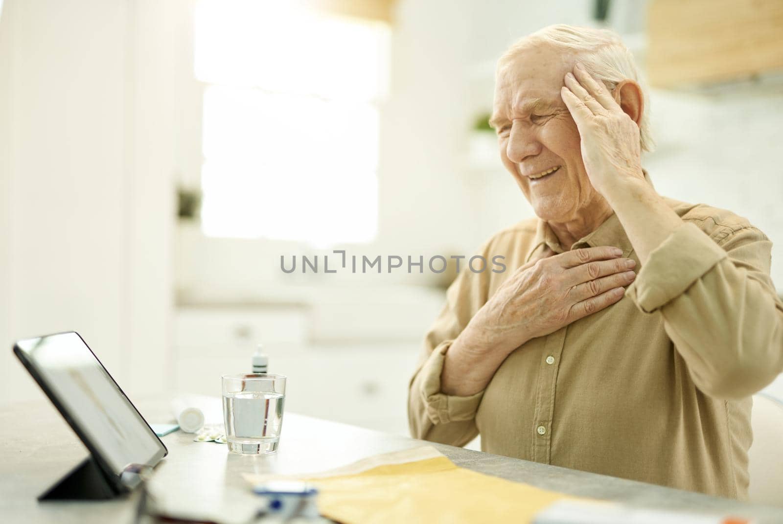 Copy-space photo of an aged man touching his head and chest while cringing and looking at the tablet screen