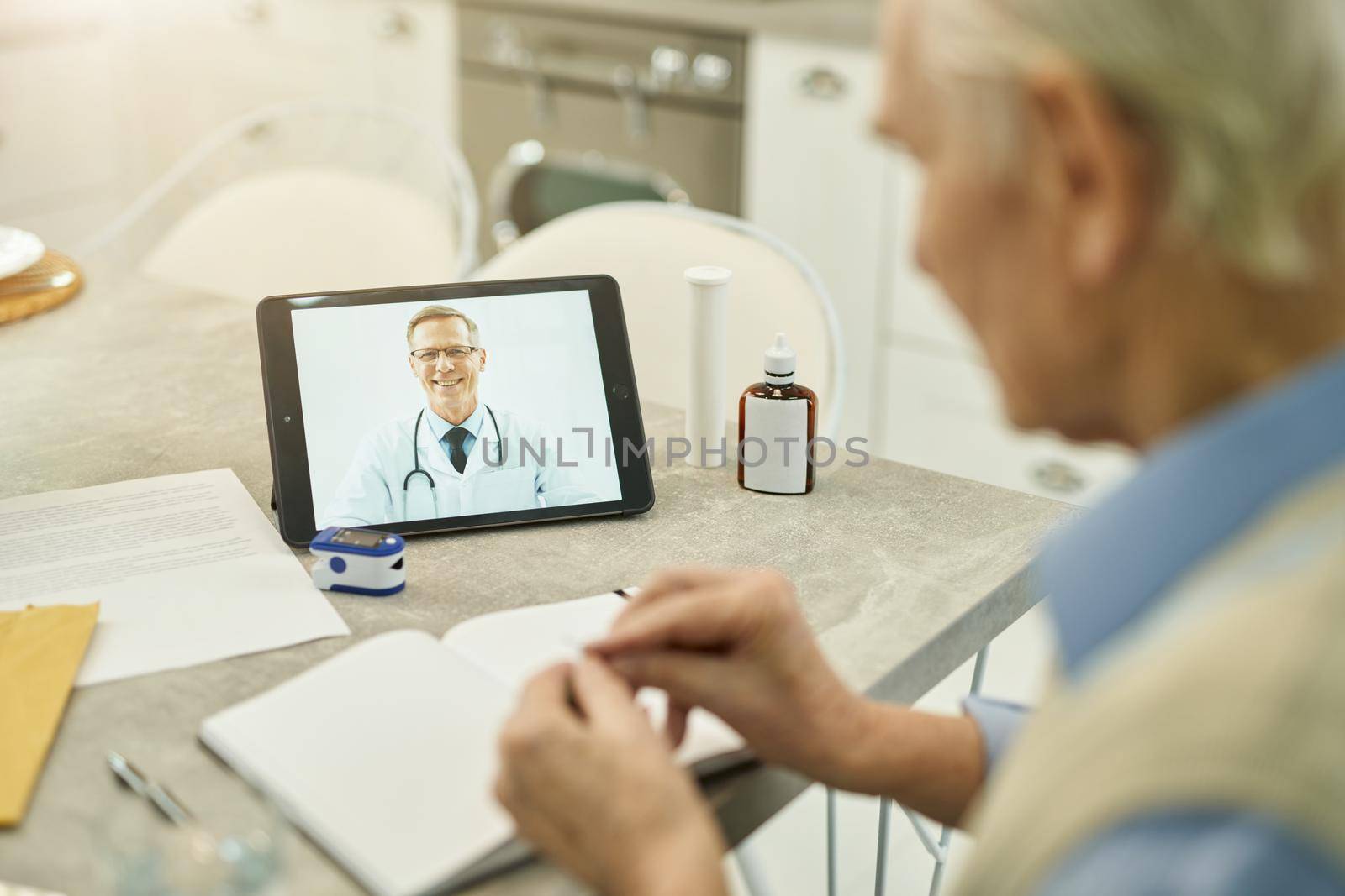 Elderly gentleman sitting at the table and looking at tablet while video-calling his healthcare provider