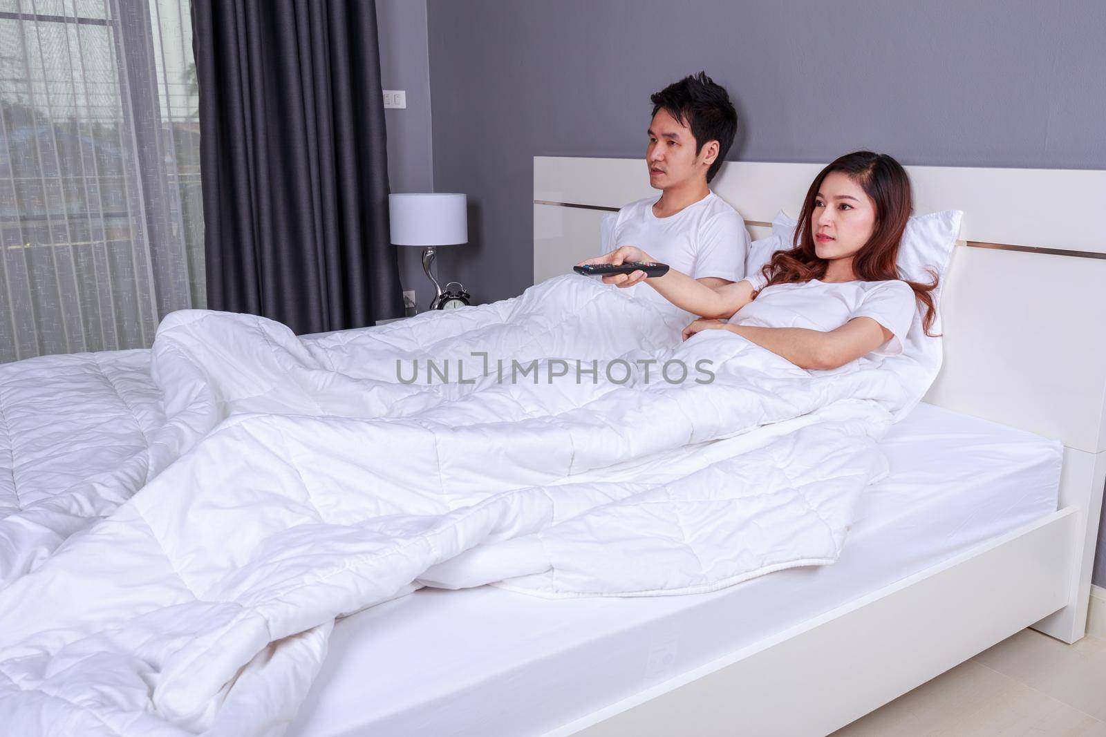 young couple lying on a bed with remote control and watching television in bedroom by geargodz