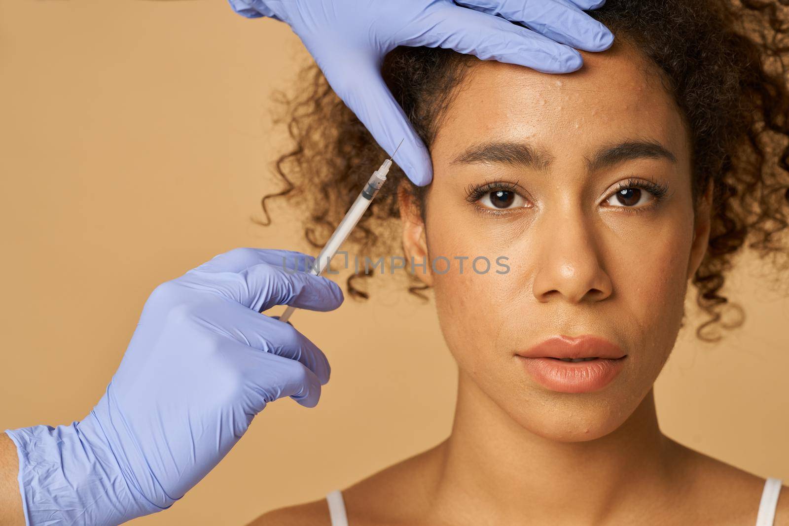 Portrait of beautiful young woman looking at camera while getting face injections isolated over beige background. Beauty, cosmetology concept