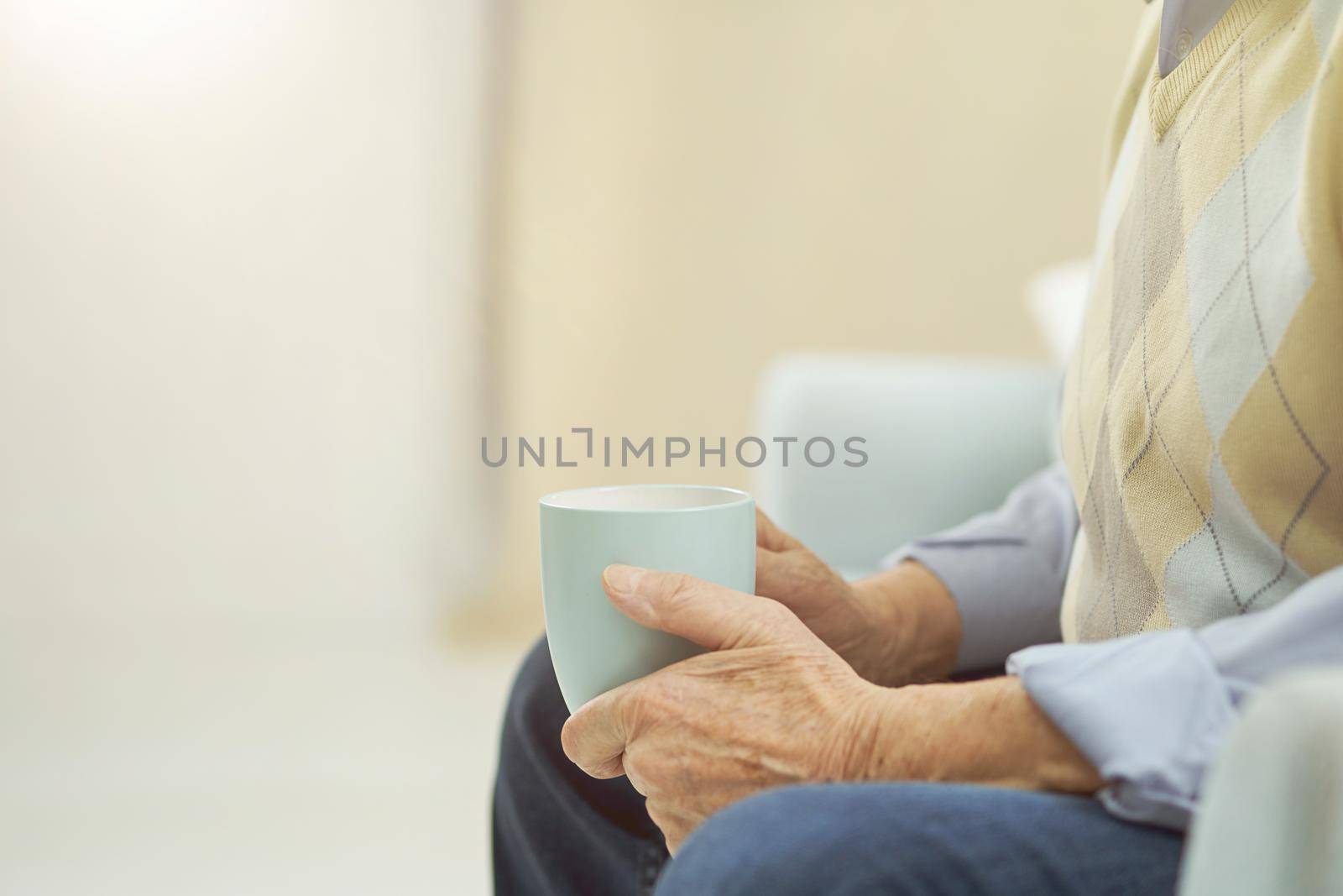 Cropped photo of elderly man sitting on the couch and holding a cup of hot drink at home. Copy space