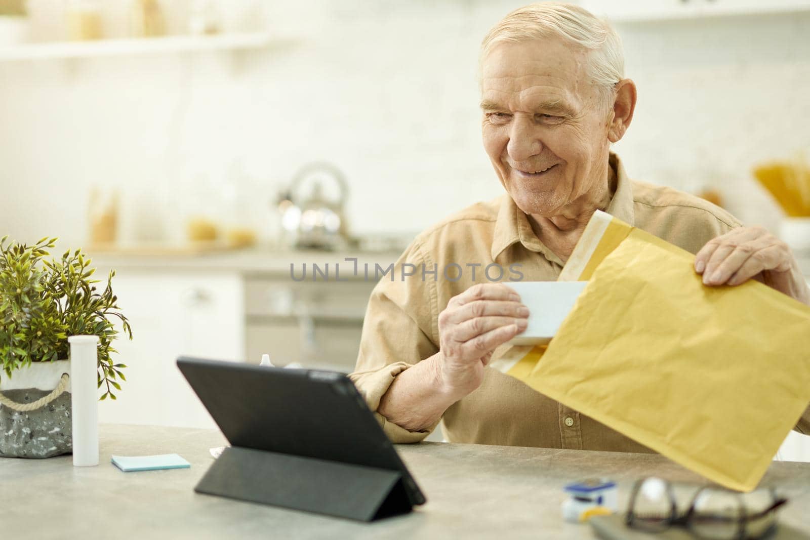 Pleased elderly man looking at tablet screen while unpacking medicine from a big postal envelope