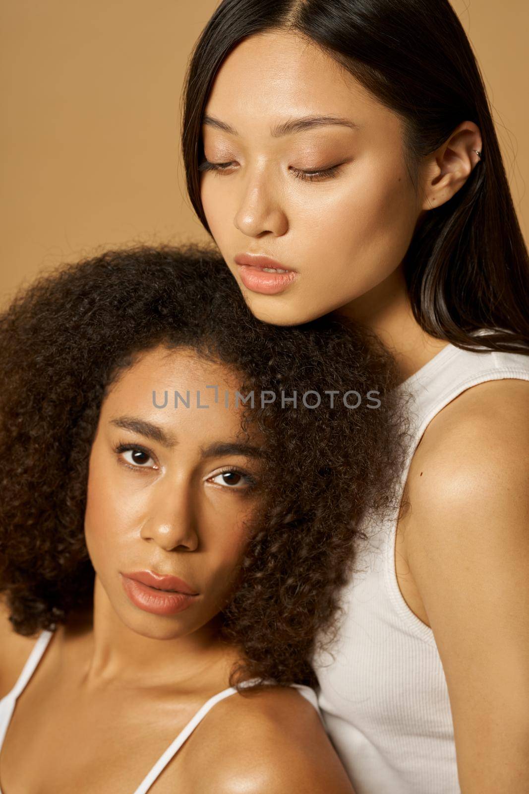 Beauty portrait of two gorgeous mixed race young women with perfect glowing skin posing together isolated over light brown background by friendsstock