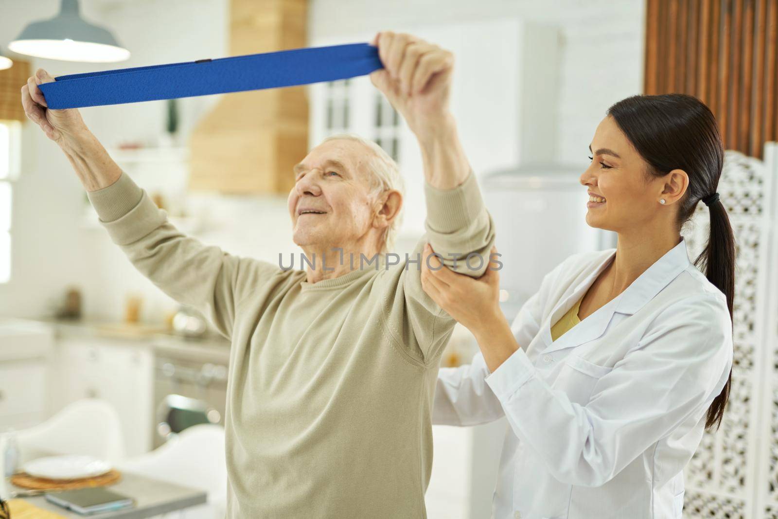 Happy senior citizen raising his hands up and doing exercises with fitness rubber band with doctor in his living room