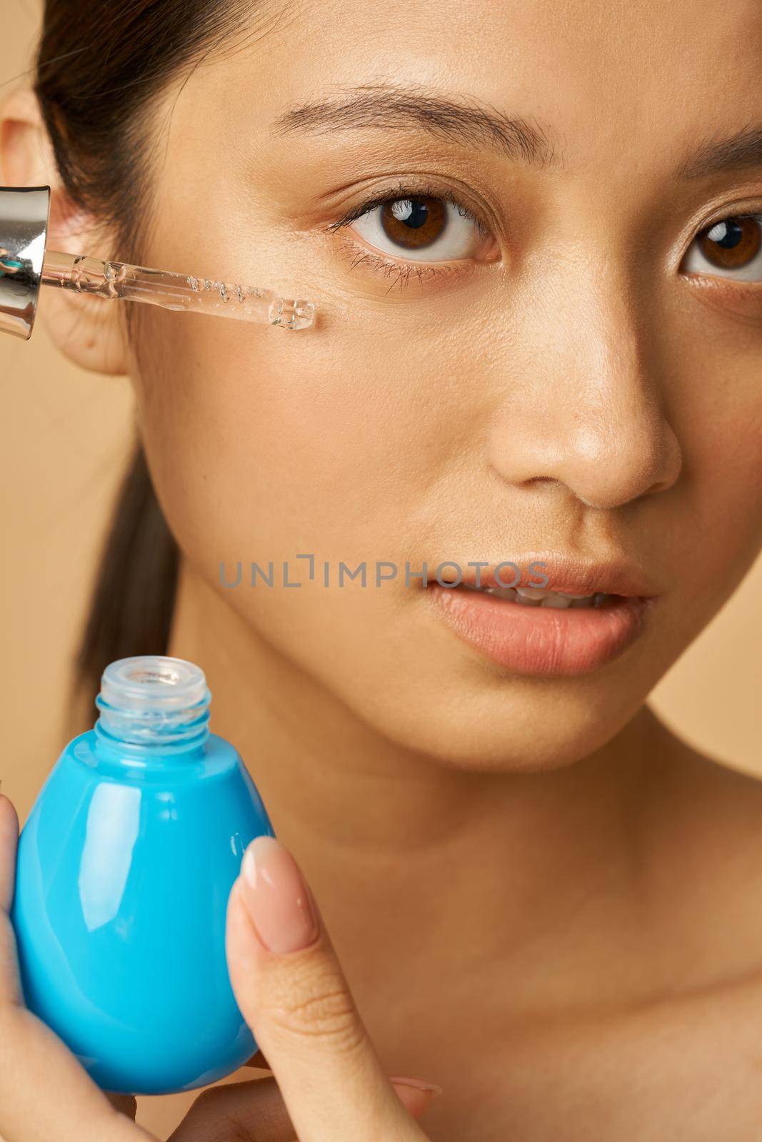 Face closeup of young woman looking at camera while applying oil drop serum, posing isolated over beige background. Beauty treatment concept