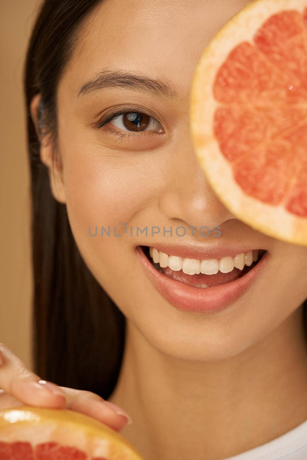 Close up portrait of joyful mixed race young woman smiling at camera, covering one eye with grapefruit cut in half, posing isolated over beige background by friendsstock