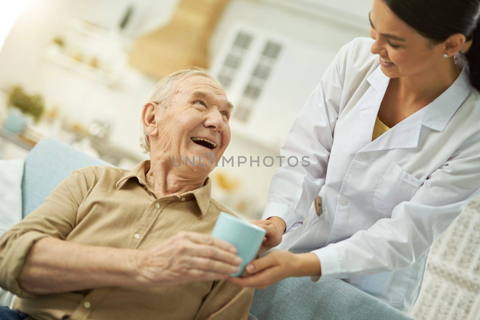 Happy young female doctor in white coat serving a cup to senior citizen at his living room while taking care of him