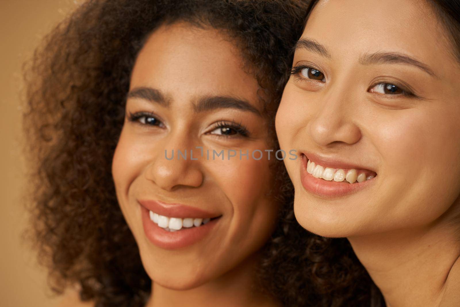 Face closeup of two joyful attractive mixed race young women with perfect smile posing for camera isolated over beige background. Health and beauty, diversity concept. Selective focus