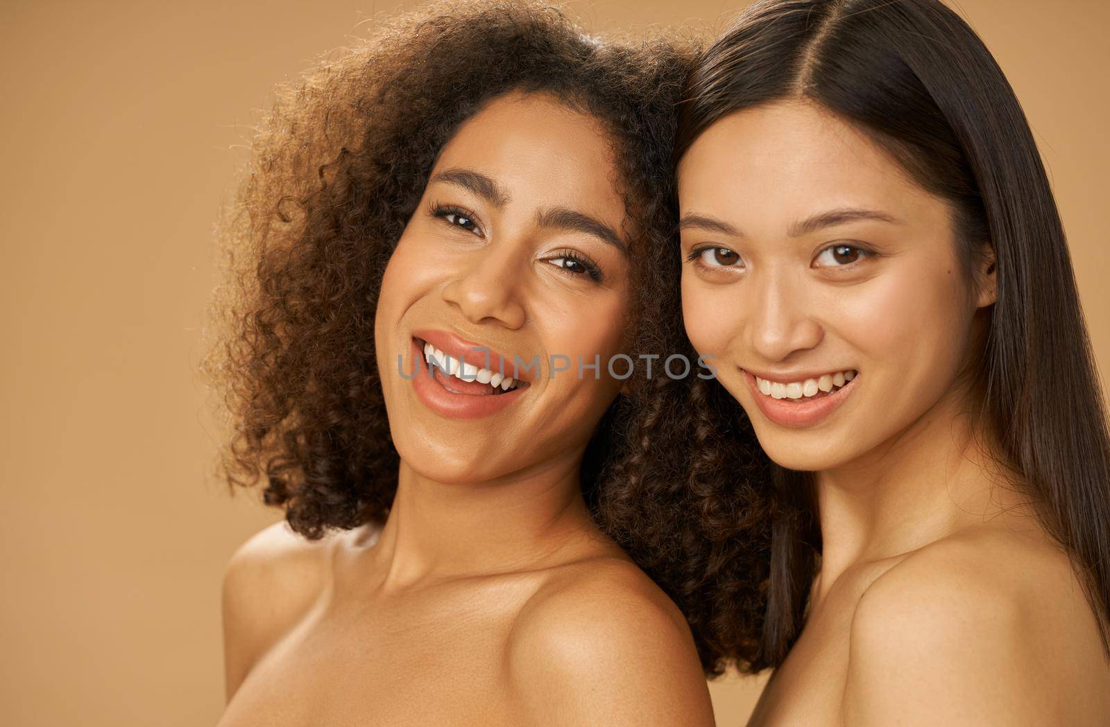 Portrait of two joyful attractive mixed race young women with perfect smile posing for camera isolated over beige background by friendsstock