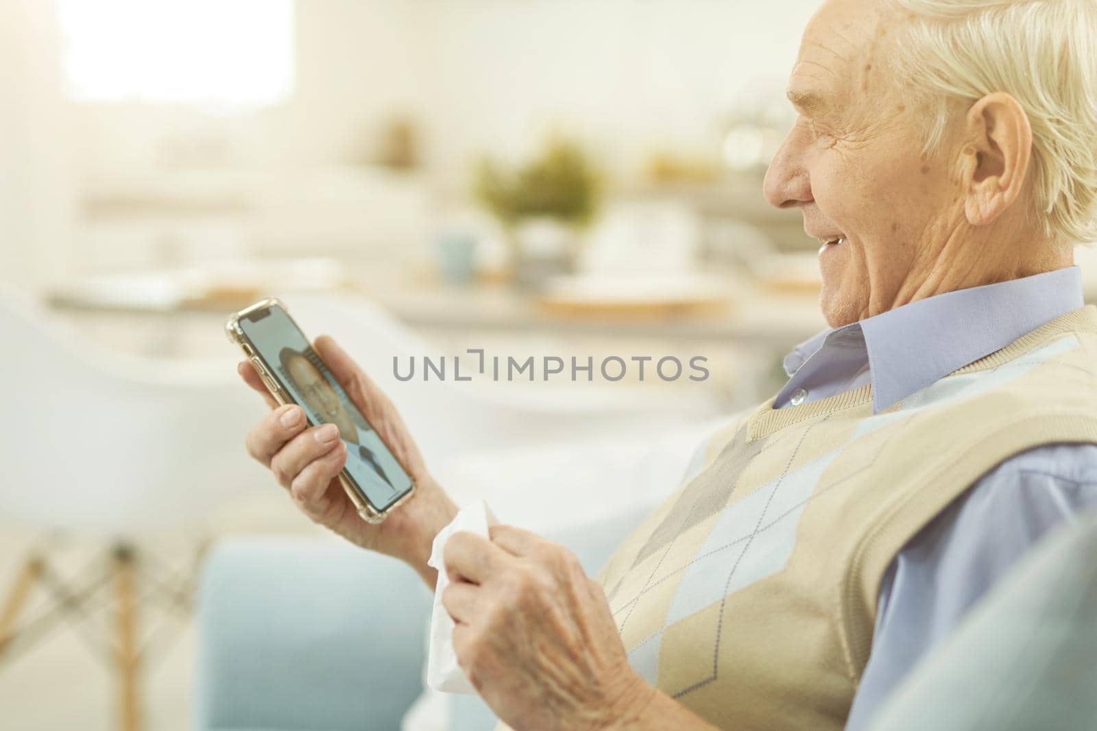 Side view of happy old man sitting at home and using mobile phone while having consultation with a therapist