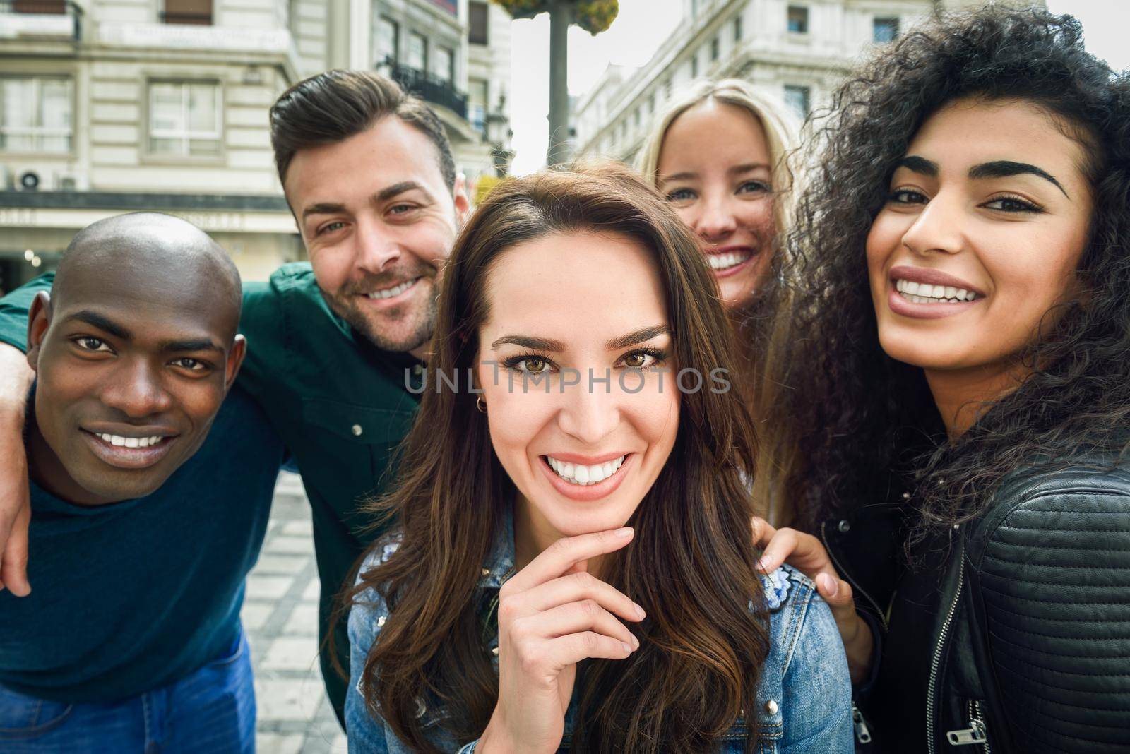 Multiracial group of friends taking selfie in a urban street with a caucasian brunette woman in foreground. Three young women and two men wearing casual clothes.