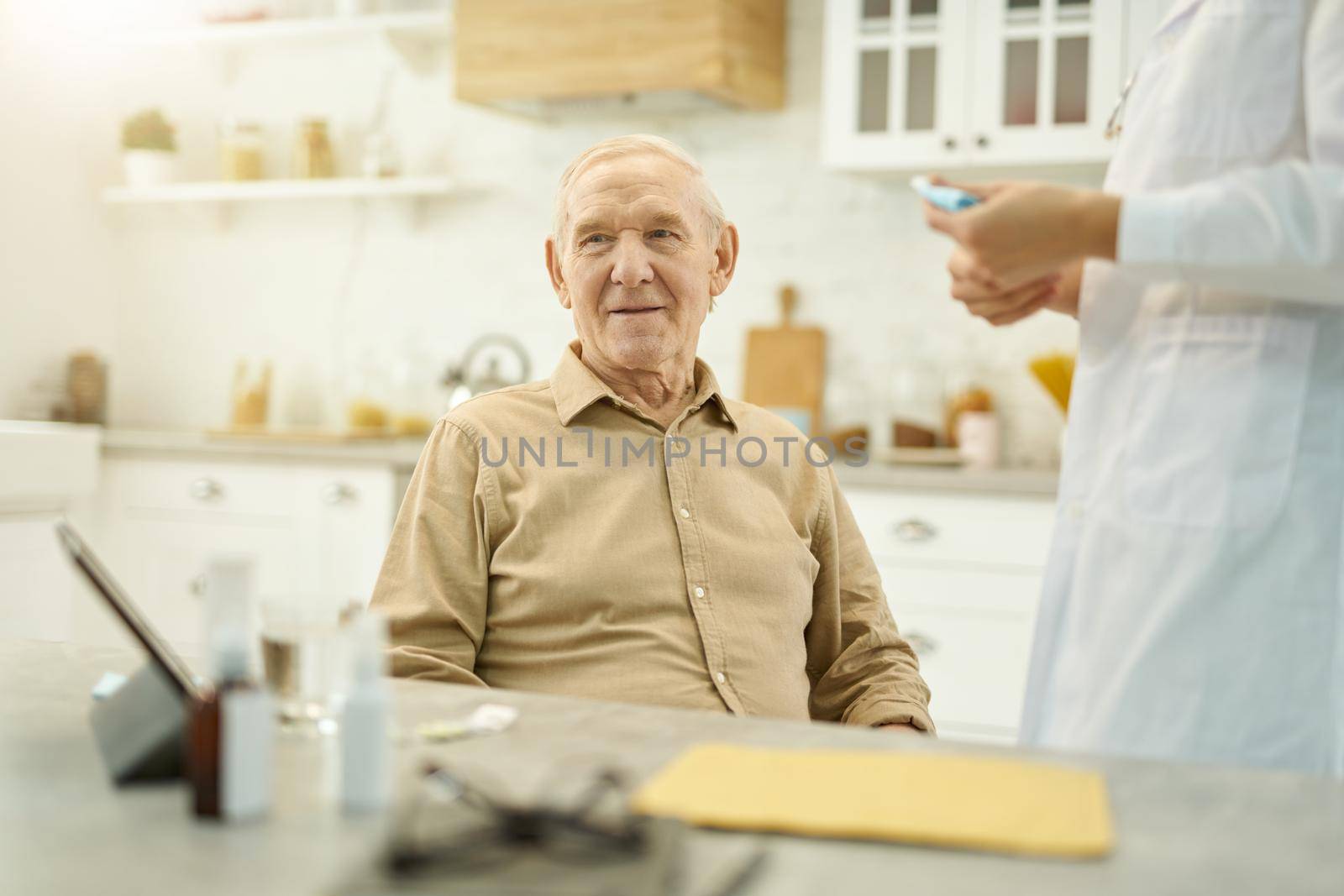 Cropped photo of a focused elderly man sitting at table while medical worker in white labcoat standing beside him
