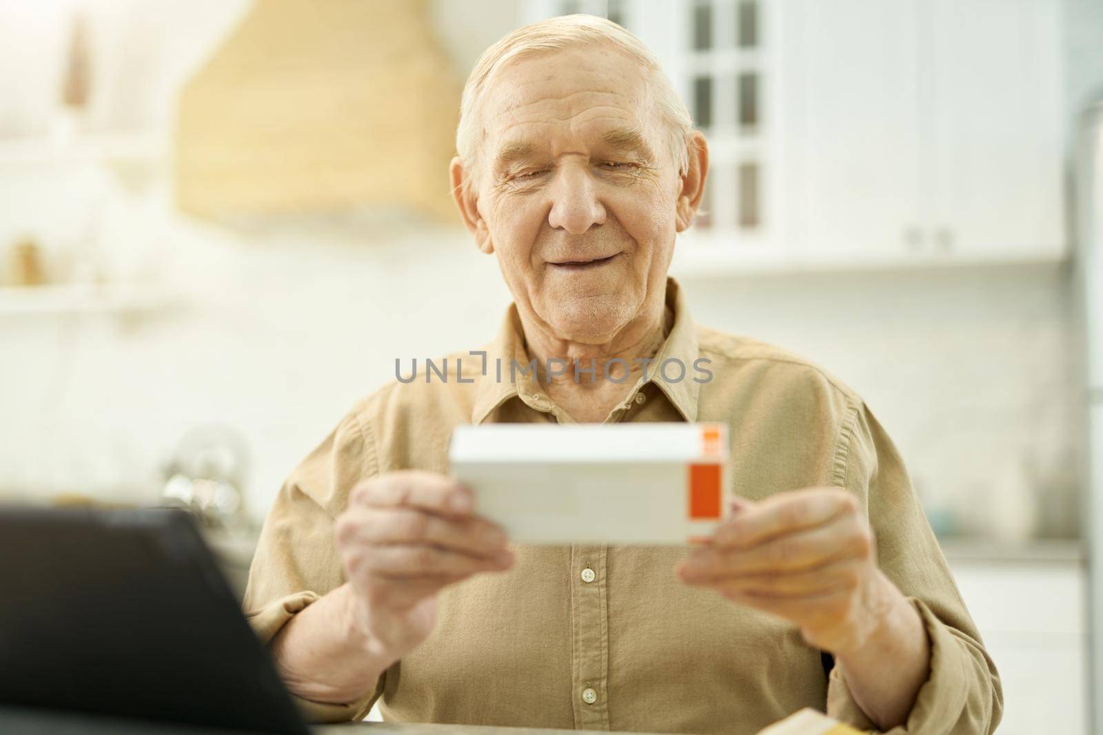 Calm senior citizen holding and looking at the tiny box of medication while sitting at the table at home