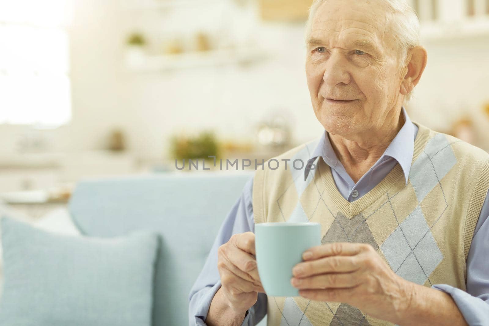 Smiling old-aged man enjoying of hot drink while sitting in living room at home. Copy space