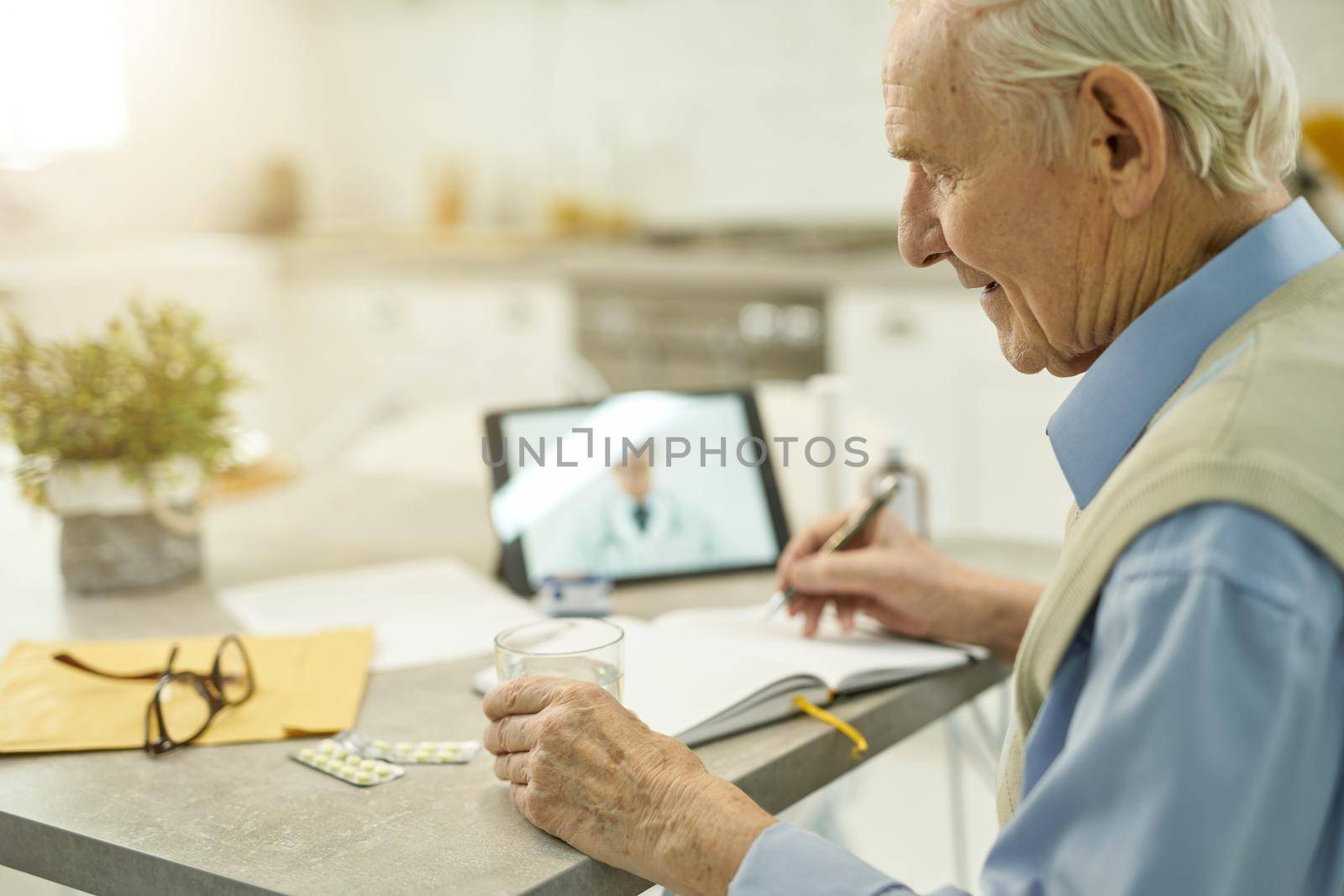Positive senior man with a pen writing down notes while having a video online consultation with his medical doctor