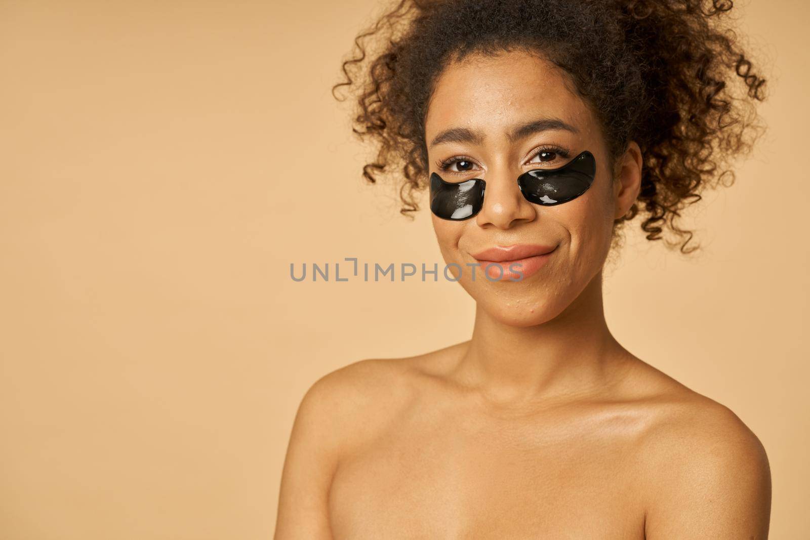 Portrait of adorable mixed race young woman posing with applied black beauty under eye patches isolated over beige background. Skincare routine concept