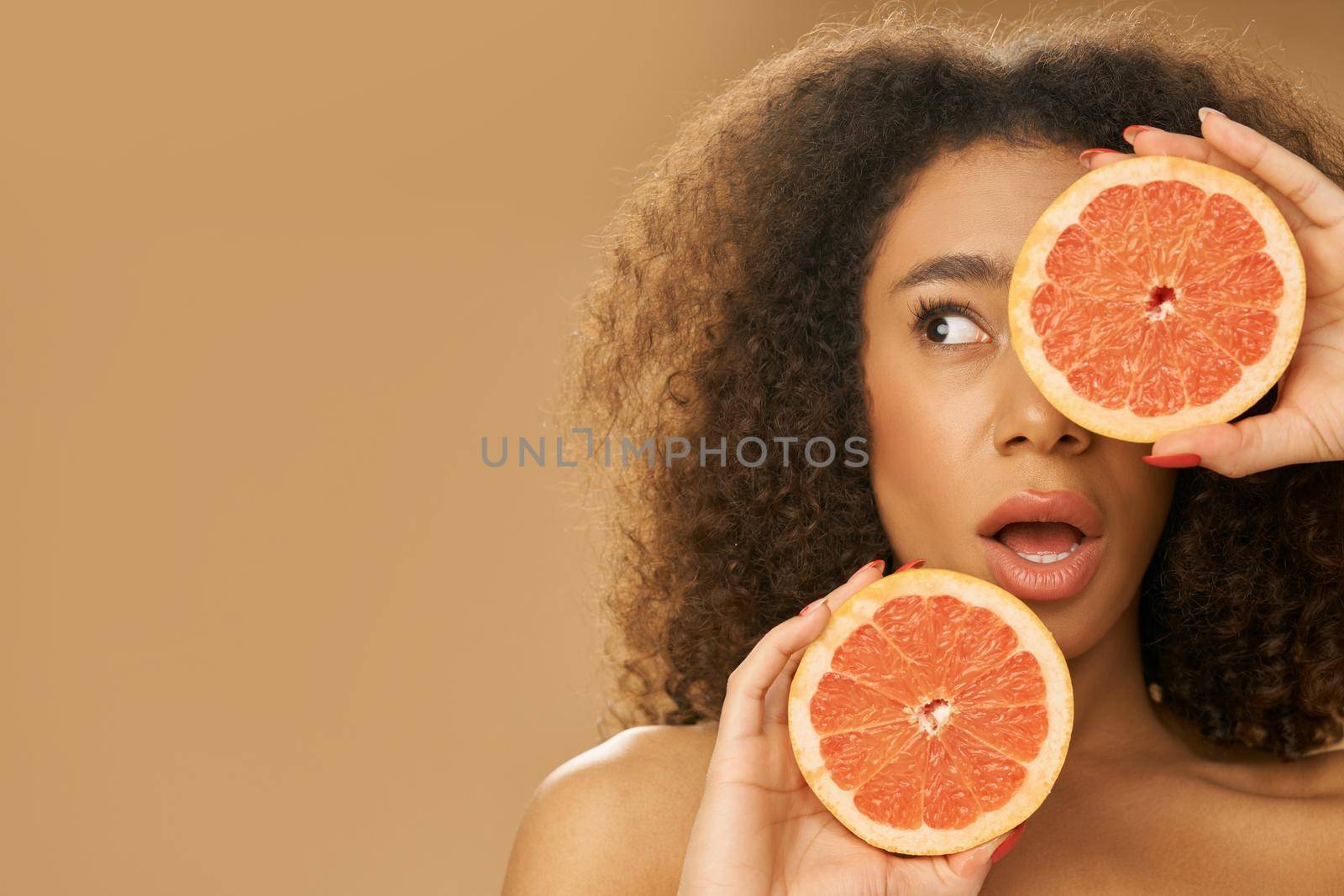 Portrait of playful mixed race young woman looking surprised, covering one eye with grapefruit cut in half while posing isolated over beige background. Health and beauty concept. Front view