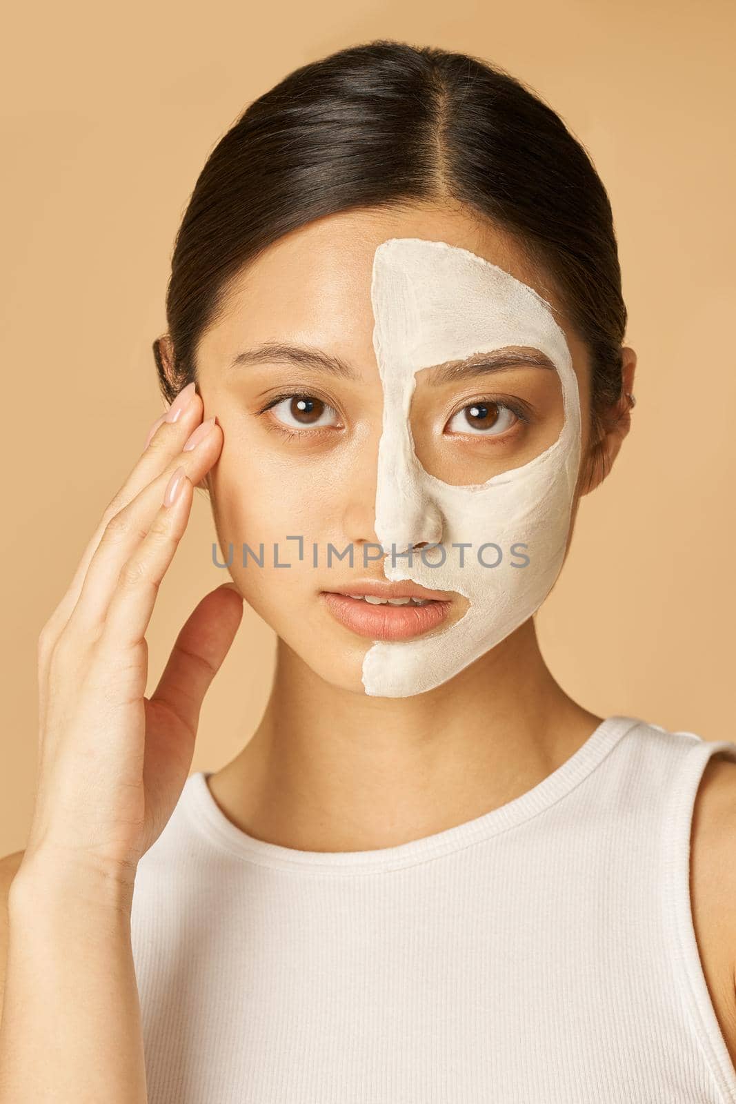 Portrait of young woman with facial mask applied on half of her face receiving spa treatments, posing isolated over beige background by friendsstock