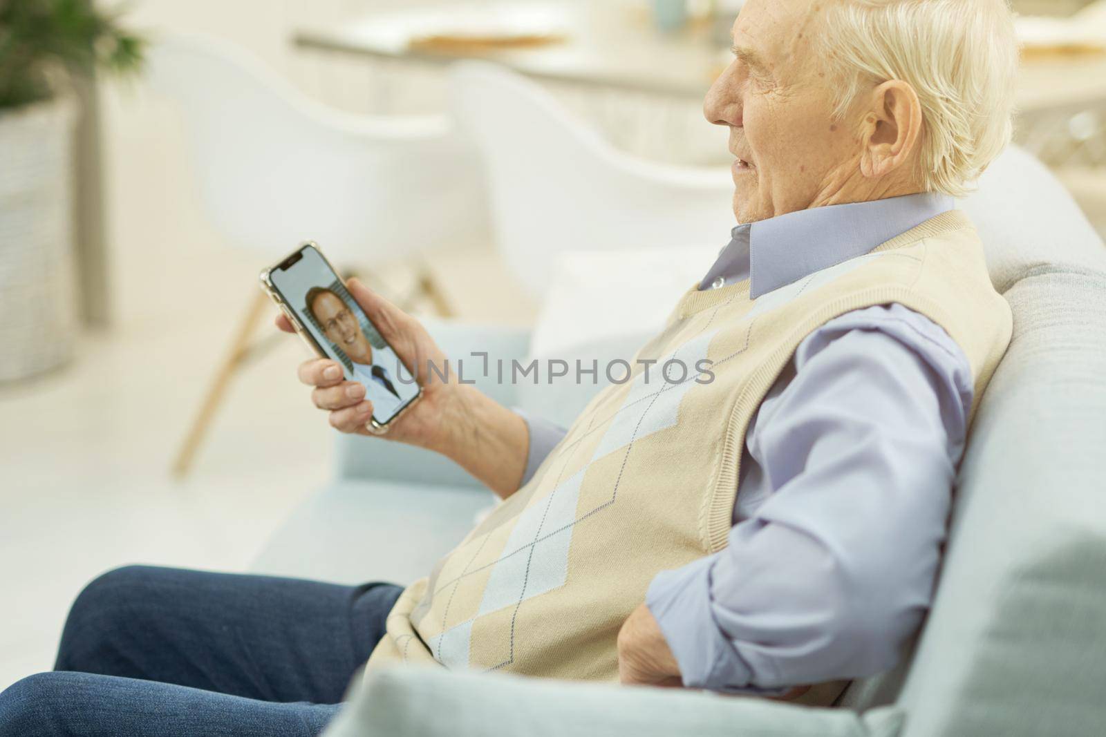 Side view of old man sitting on sofa at home and using mobile phone during a consultation with a therapist