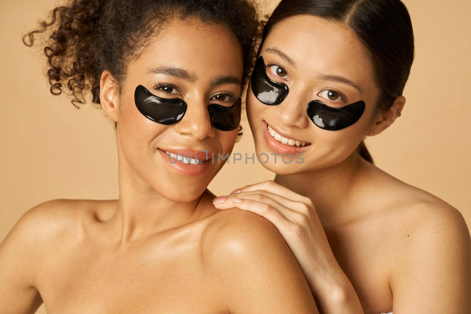 Beauty portrait of two adorable young women posing with applied black under eye patches, standing together isolated over beige background. Skincare routine concept