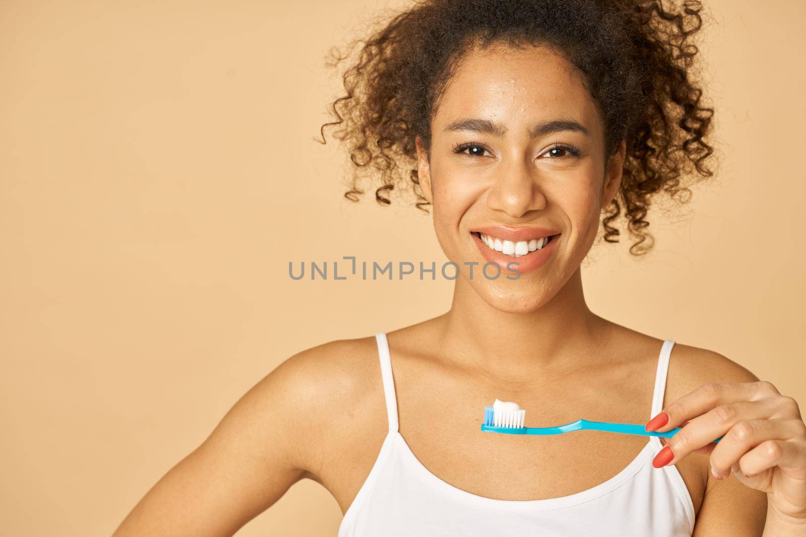 Portrait of cheerful young mixed race woman brushing teeth in the morning, posing isolated over beige background by friendsstock