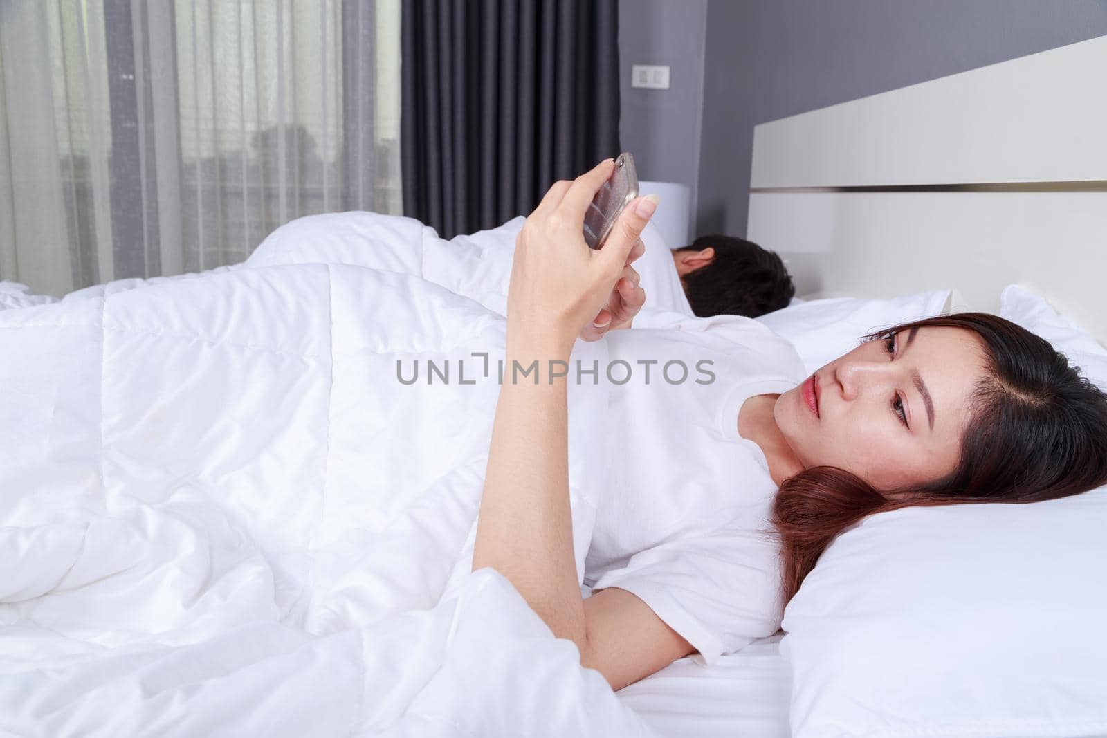 young woman using her mobile phone in bed while her husband is sleeping next to her
