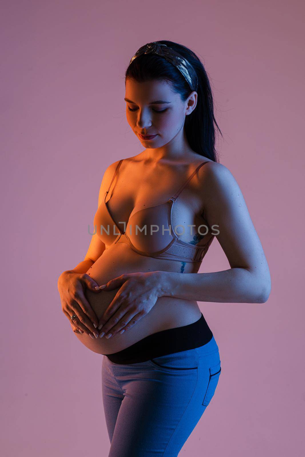 Studio portrait of attractive slim pregnant woman with dark hair with band standing in bra and jeans holding heart gesture with hands on tummy. Isolate on gentle pink background.