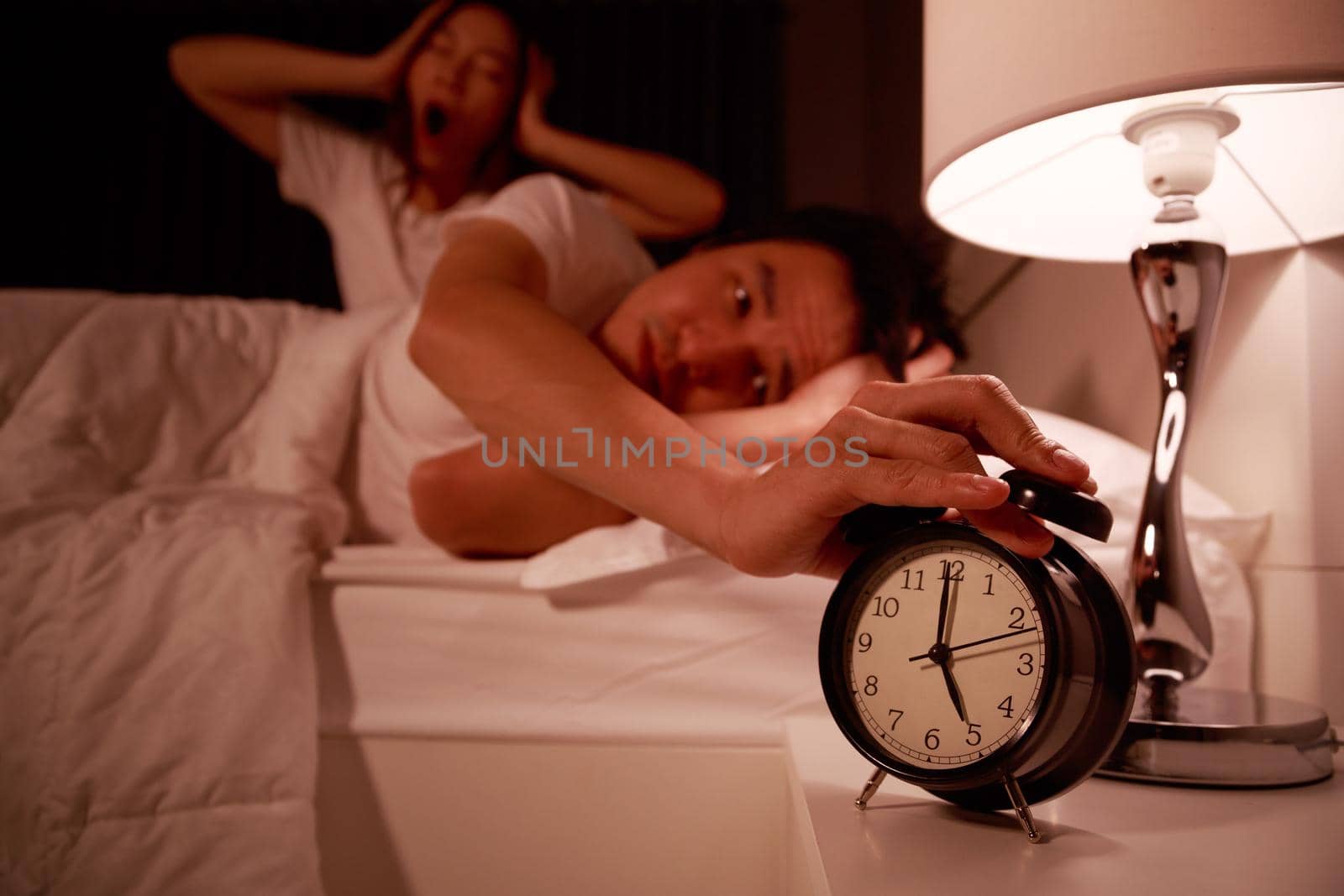 sleepy couple in bed with extending hand to alarm clock in the morning by geargodz