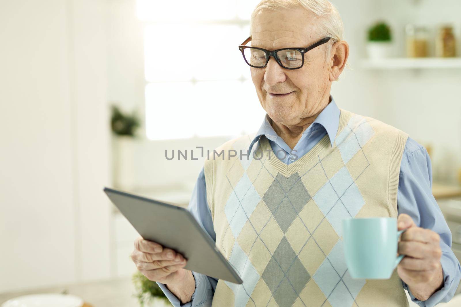 Elderly man in glasses holding a cup of tea while looking at the tablet screen and making a video call