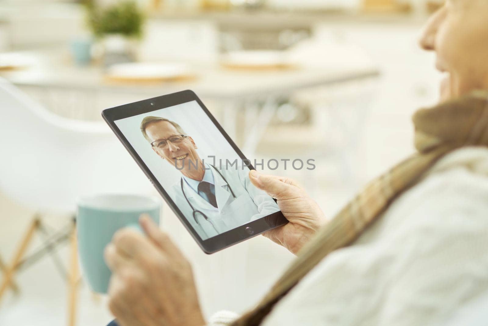 Elderly sick male holding a cup of drink while looking at the tablet screen and making a video call