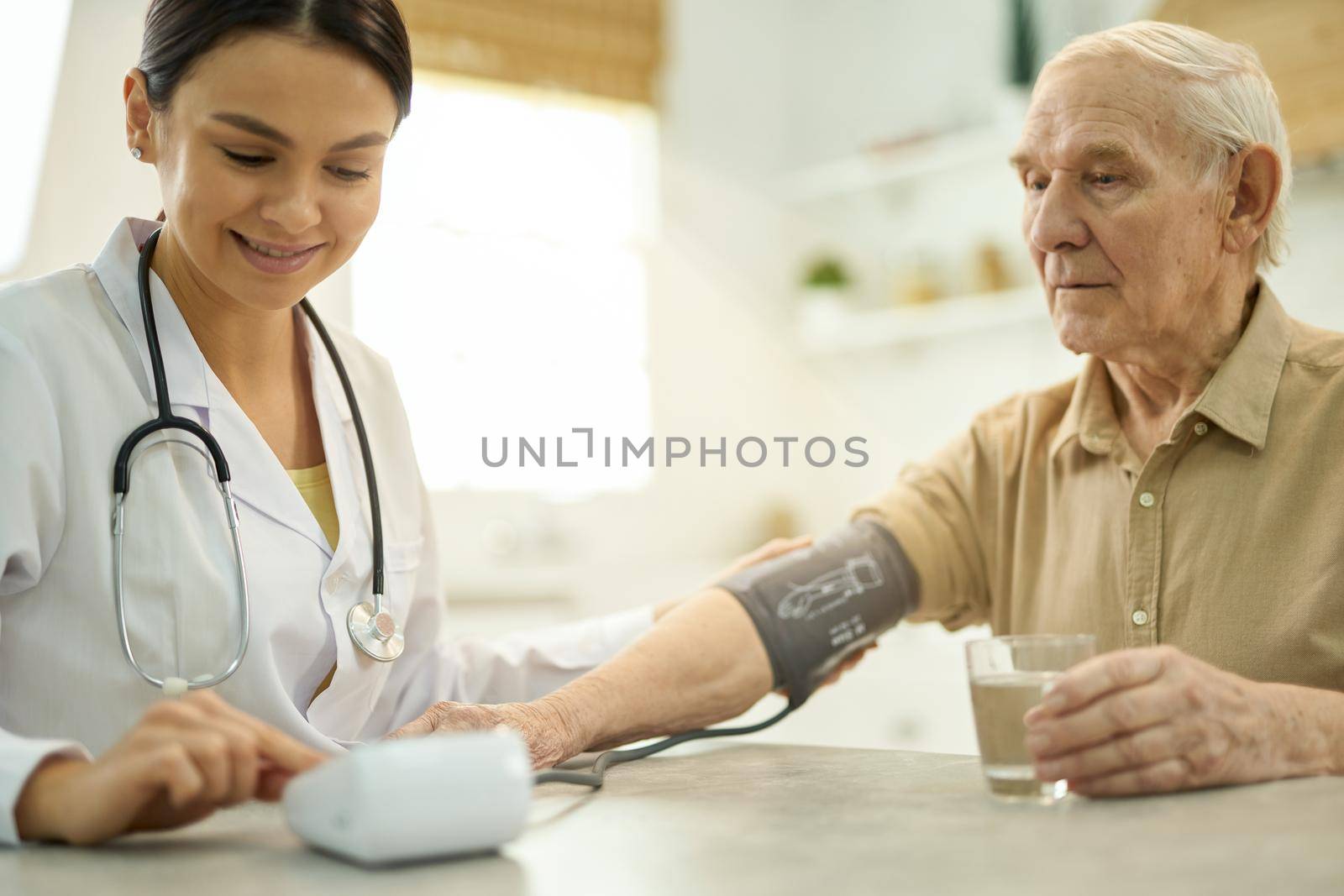 Friendly female medic using a manometer to measure serious senior man blood pressure while sitting at the table