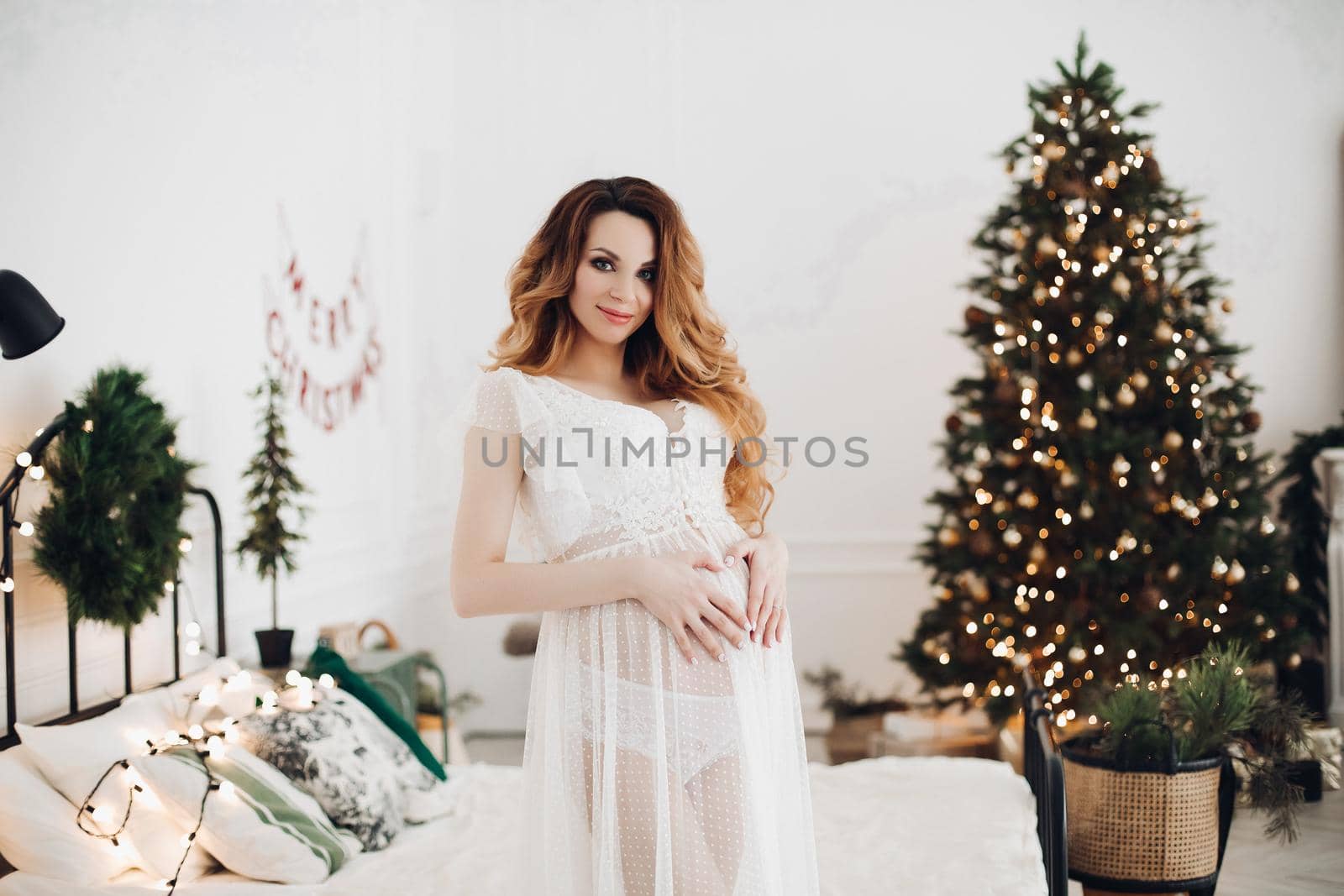 Portrait of smiling pregnant young woman tenderness holding big tummy showing heart medium shot. Beautiful girl making love gesturing posing enjoying pregnancy at Christmas tree background