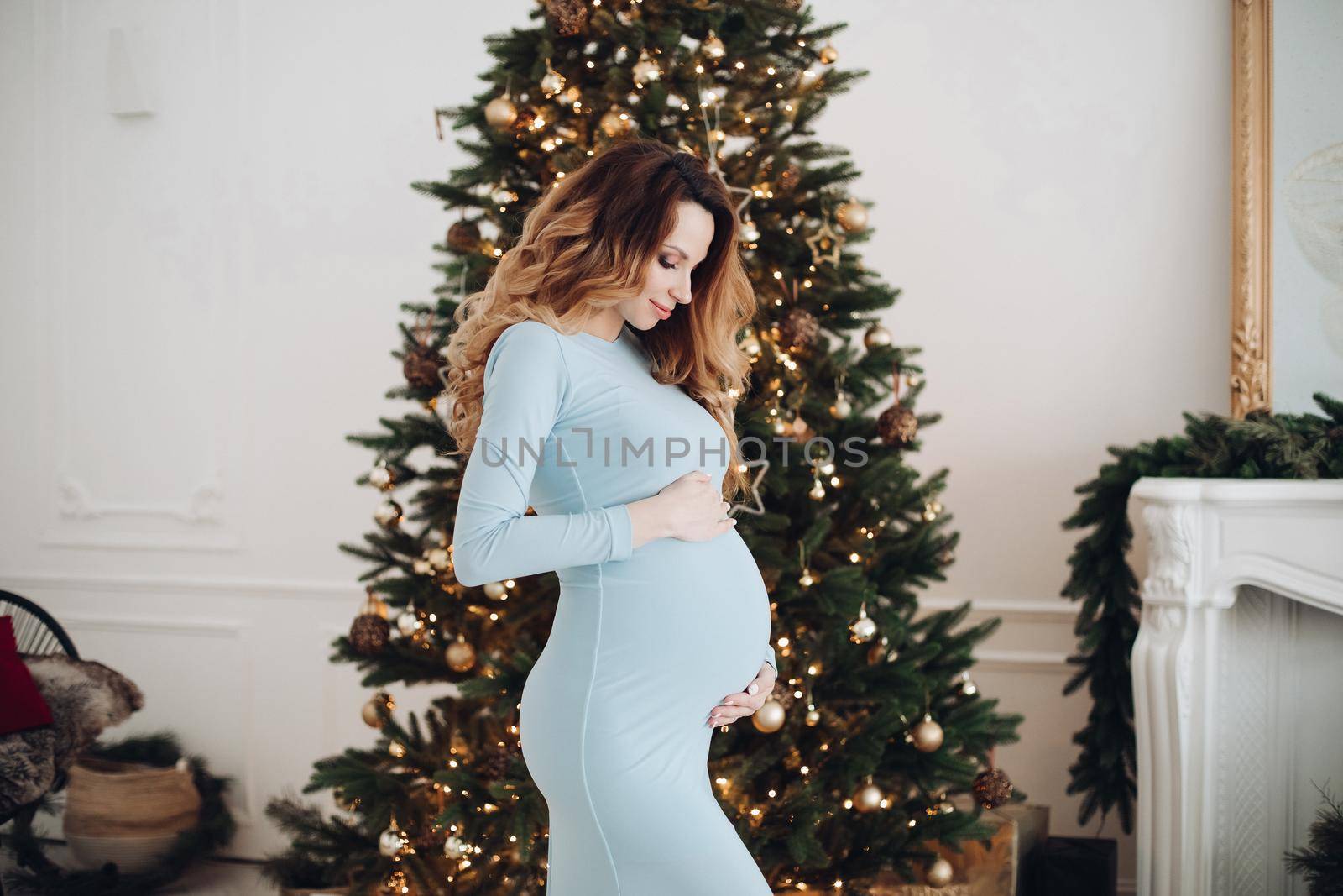 Full length of pregnant female in blue dress smiling joyfully in studio on the background of New Year lights. Christmas and New Year concept