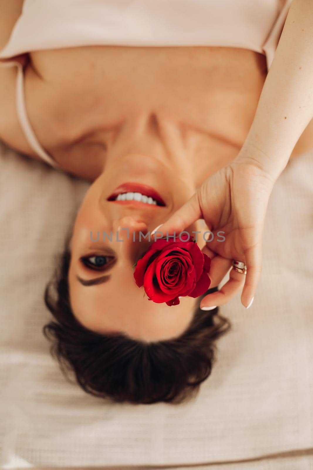 Overhead portrait of happy smiling woman with dark-hair laying on bed and holding red rosebud in front of her eye, looking at camera.