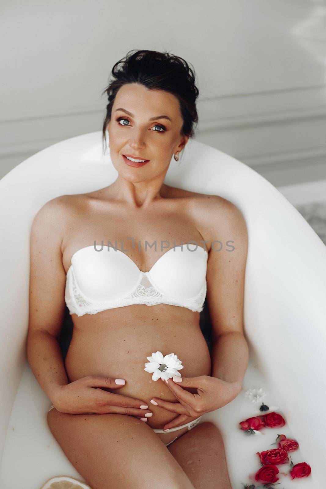 Portrait of attractive pregnant adult brunette in white bra laying in bath with white flower on belly button and red roses in water. Pregnant woman smiling at camera.