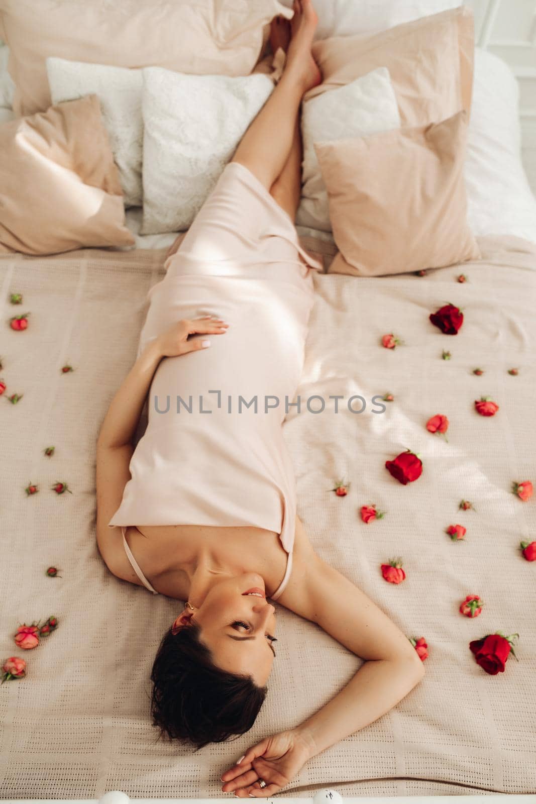 Upside-down shot of a relaxed brunette woman in beige dress laying on a bed sprinkled with flower petals