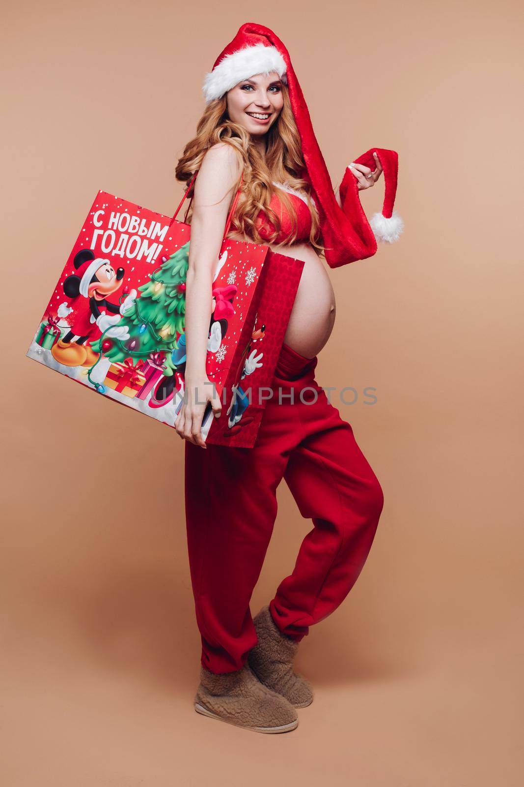 11 27 2019 Belarus Minsk: Waist up of pretty pregnant lady touching Santa Claus hat while loking at camera on beige background. Christmas and New Year concept