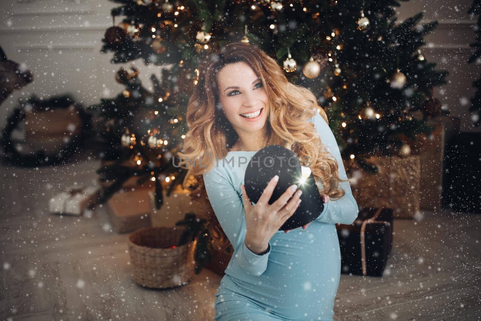 Waist up of smiling pretty pregnant lady opening present while sitting near Christmas tree. Christmas and New Year concept