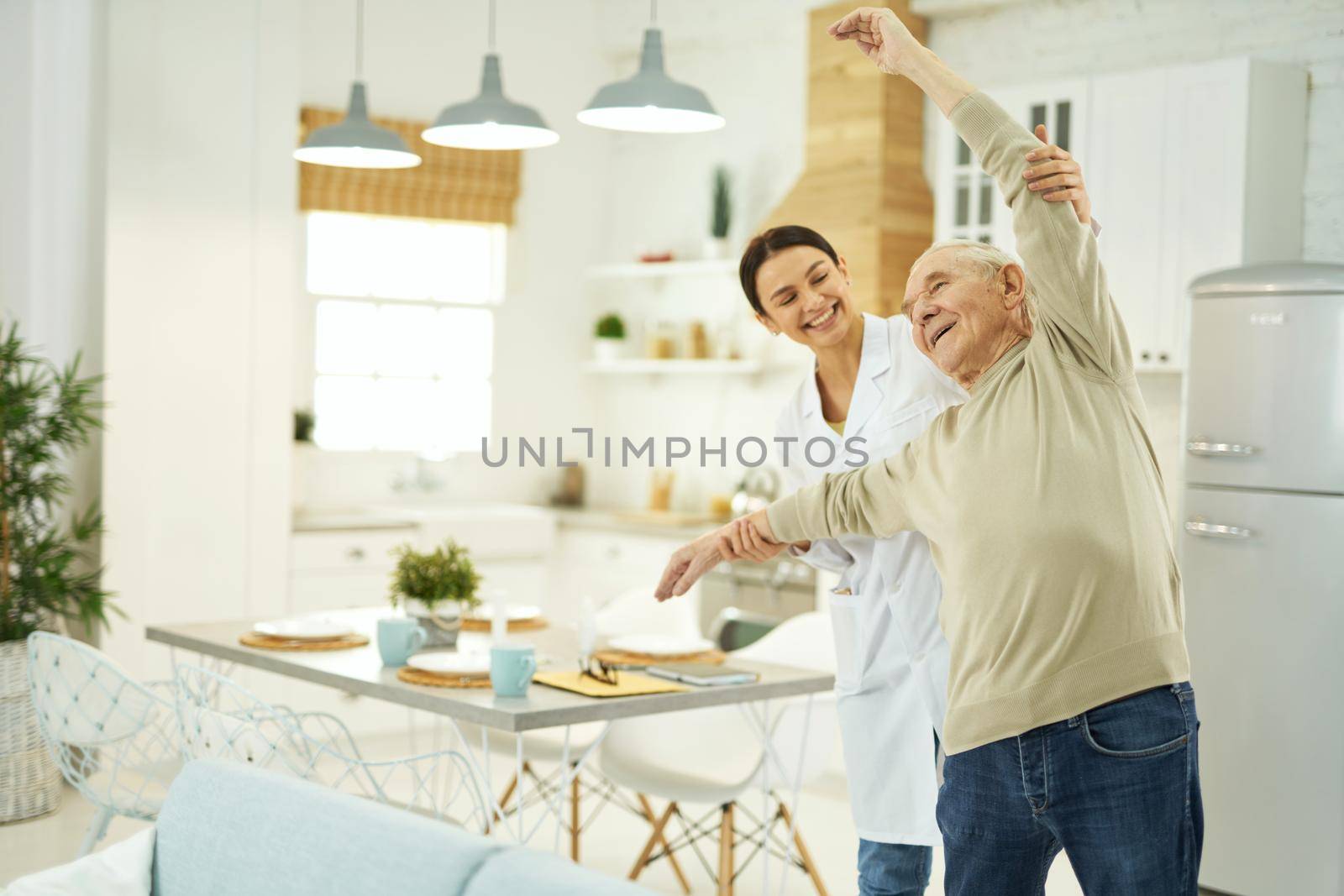 Smiling young female doctor in white coat helping an elderly man to do exercises in his apartment