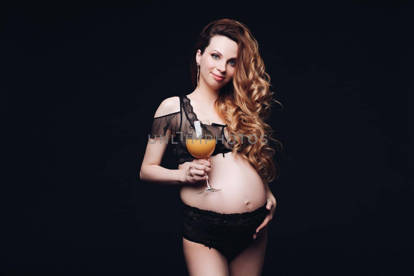 Portrait of happy beautiful pregnant young girl with bocal of fresh orange juice medium long shot. Smiling attractive pregnancy female posing at black studio background enjoying healthy lifestyle