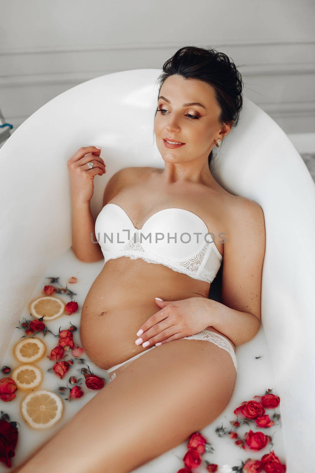 Lovely pregnant lady in a bikini relaxing in a soothing bath-tub with flower petals and orange petals