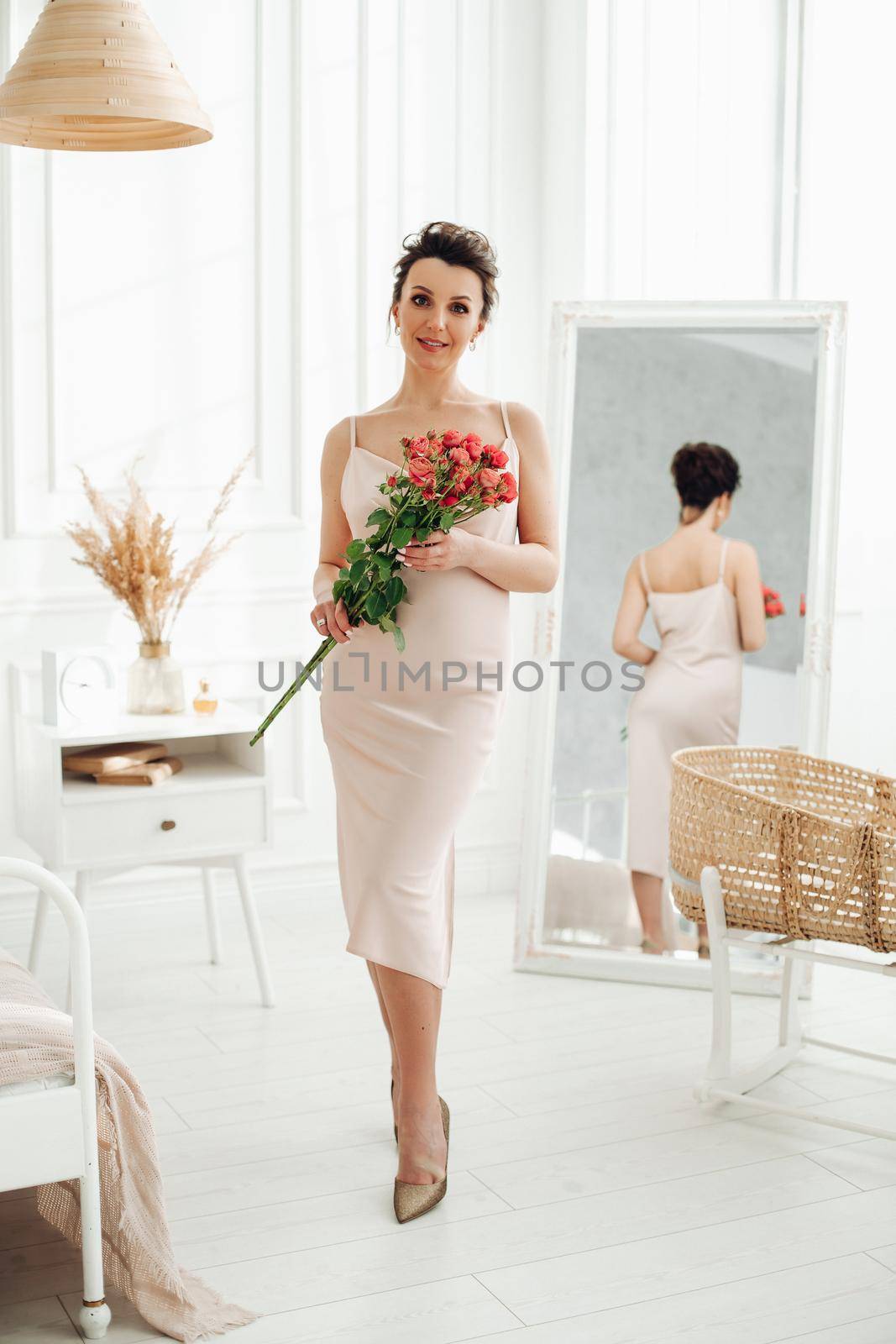 Full length portrait of pregnant gorgeous dark-haired Caucasian woman in beige silk dress and heels holding bouquet of red roses and smiling at camera. Mirror reflection of her in the background.